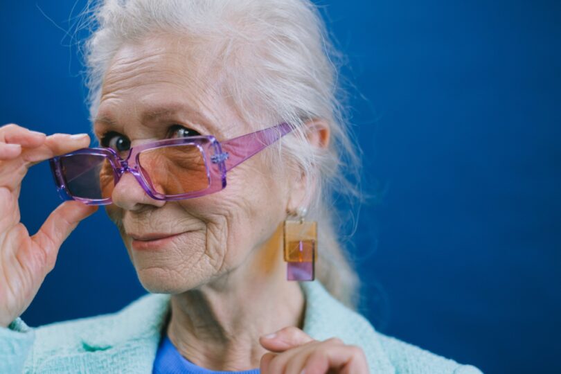 Portrait of elegant smiling gray haired elderly female wearing purple sunglasses and earrings looking at camera against blue background